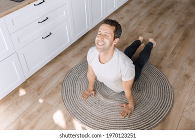 Motivated Joyful Guy Doing Yoga Exercises During Home Training. Smiling Man Doing Cobra Pose At Carpet On The Floor In Modern Kitchen, Relieve Stress Doing Physical Exercises