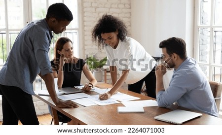 Motivated millennial African American workers discussing marketing strategy, analyzing sales paper report with skilled Caucasian colleagues at briefing meeting in modern office, collaboration concept.