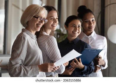 Motivated Multiracial Female Team Office Workers Of Different Generations Look At One Side Smile Ready To Meet Help Client. Professional Diverse Businesswomen Stand Together Hold Documents Clipboards