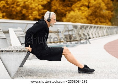 Motivated sporty elderly man in black sportswear have workout outdoor at autumn park, standing by bench, stretching body, sportsman using wireless headphones, listening to music, copy space