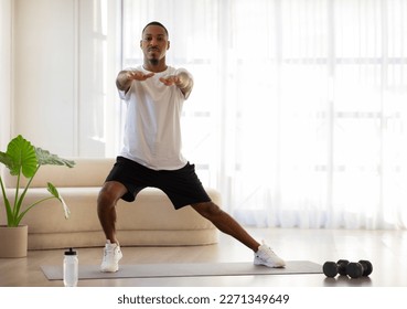 Motivated sporty handsome young black man in sportswear standing on yoga mat, doing squats, have workout at home, copy space. Fitness at home concept - Powered by Shutterstock