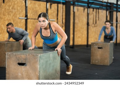 Motivated sporty young girl performing incline push-ups on plyometric box, focused on chest, shoulders, triceps, and abs training during intense group workout at gym - Powered by Shutterstock