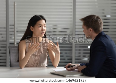 Motivated young Asian female work candidate talk share thoughts with male employer or recruiter, serious millennial biracial woman applicant speak with hr manager at job interview in office