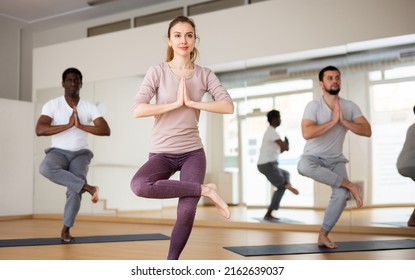 Motivated young attractive woman standing in balancing asana Eka Pada Utkatasana with hands clasped in prayer during group yoga class in fitness studio