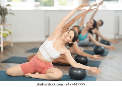 Motivated young girl performing Mermaid Stretch with Ball in group Pilates class, focusing on flexibility and core strength, in light-filled airy studio - Powered by Shutterstock