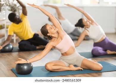 Motivated young girl performing Mermaid Stretch with Ball in group Pilates class, focusing on flexibility and core strength, in light-filled airy studio - Powered by Shutterstock