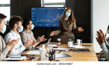 Motivation Speech. Smiling Young Female Ceo In Medical Protective Mask Making Presentation To Audience During Meeting With Employees, Standing At Table In Board Room. People Clapping Hands