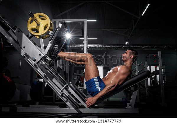 Motivational portrait of handsome muscular\
brutal European man in shorts working on legs press machine in\
modern gym. Night workout\
concept.