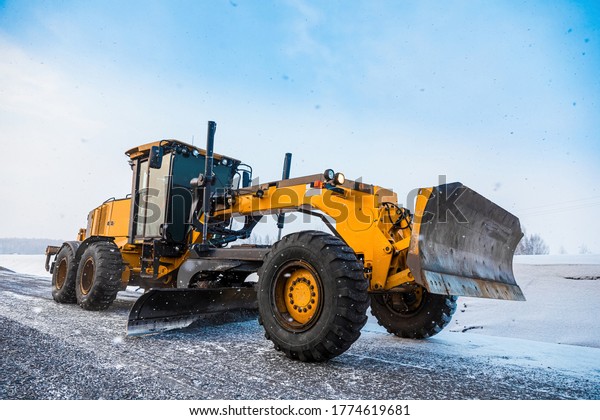 Motor Graders, Grader\
Road Construction. Industrial machine on construction of roads.\
Truck moving on dirt country road. Grader is driving along an empty\
winter road