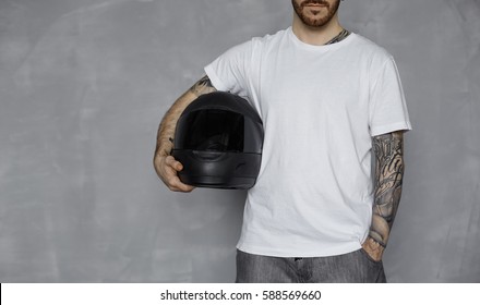 Motorbiker Concept With Blank White T-shirt. Brutal Bearded Man With Tattooed Arms Keep Black Motorcycle Helmet On Neutral Grey Background In Studio. Extreme Rider Life