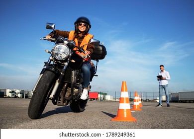Motorcycle school of driving. Female driver with helmet taking motorcycle lessons and practicing ride. In background traffic cones and instructor with checklist rating and evaluating the ride. 