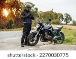 motorcyclist in a helmet and motorcycle clothing stands at an old cafe racer with a smartphone
