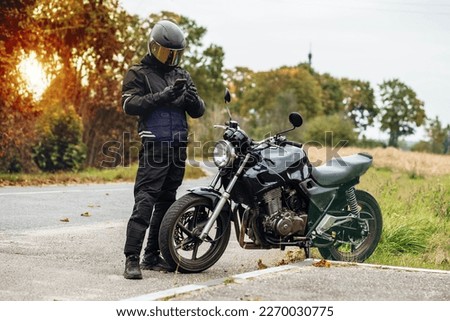 motorcyclist in a helmet and motorcycle clothing stands at an old cafe racer with a smartphone