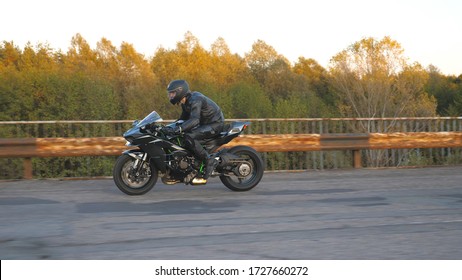 Motorcyclist In Helmet Riding Fast On Modern Sport Motorbike At Old Rusty Bridge On Highway. Young Man Racing His Motorcycle On Country Road. Guy Driving Bike. Concept Of Post Apocalypse. Side View.