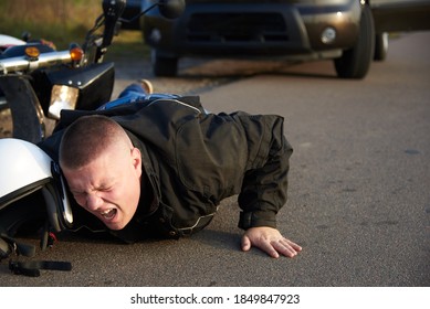 A Motorcyclist Lies On The Asphalt Near A Motorcycle And Car, The Theme Of Road Accidents.