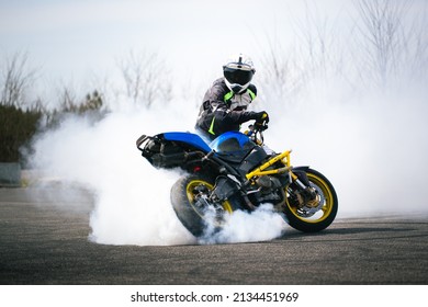 A motorcyclist in protective gear executes a dramatic burnout on a paved track, creating smoke and dynamic motion. The blue and yellow sport bike adds color and energy to the scene.