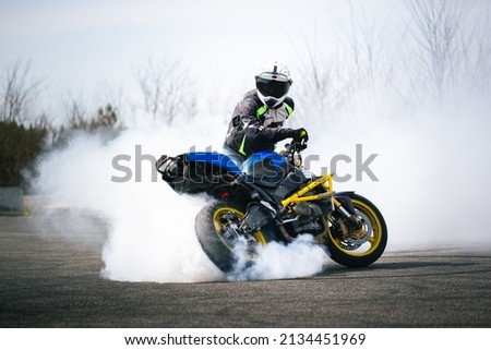 A motorcyclist in protective gear executes a dramatic burnout on a paved track, creating smoke and dynamic motion. The blue and yellow sport bike adds color and energy to the scene.
