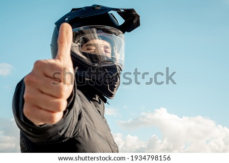 Motorcyclist wearing a black dual-purpose motorcycle helmet with a black jacket with sky background and thumbs up.