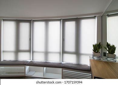 Motorized roller shades in the interior. Automatic roller blinds beige color on big glass windows. Home luxury curtaines are above the windosill with pillows. Summer. Green trees outside.