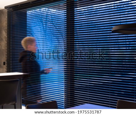Motorized wood blinds black color in the interior. Automatic venetian blinds closeup on the large windows. The park is visible through the blinds. The girl holds a blinds control panel in her hands. 