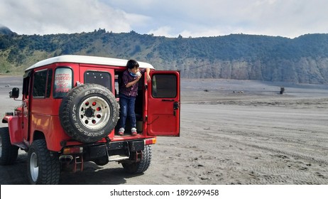 Mount Bromo, Indonesia - Dec 2020: Close up of young boy standing on the back of red Japanese classic jeep in the desert with cloudy sky background.