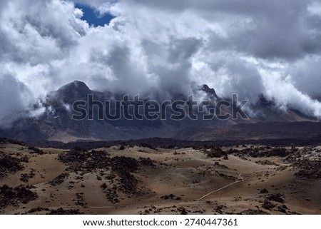 Mount Guajara rises dramatically through swirling low clouds above the volcanic pumice mounds and lava fields of the Las Cañadas caldera floor, photographed with a telephoto compression that emphasises the imposing scale of the mountain, Teide plateau, Tenerife