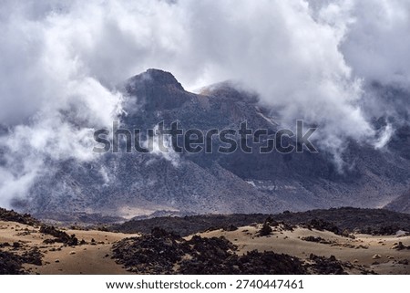 Mount Guajara rises dramatically through swirling low clouds above the volcanic pumice mounds and lava fields of the Las Cañadas caldera floor, photographed with a telephoto compression that emphasises the imposing scale of the mountain, Teide plateau, Tenerife
