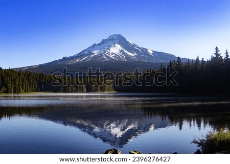 Mount Hood reflecting in Oregon's Trillium Lake early morning with steam on water bordered by lush pine forest.