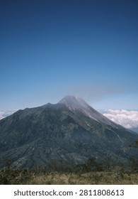 Mount merapi in indonesia, an active stratovolcano with green slopes and rugged peaks under a blue sky with clouds. Ideal for travel websites and geological studies.