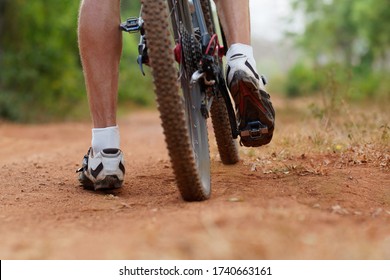 Mountain Bike Rear Wheel And Riders Foot. Back Shot Of Mountain Bike On Brown Dirt Road. Close Up View Of A Mountain Bike Tire.
