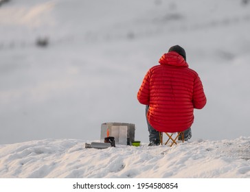 Mountain Climber In Red Coat On White Snow Covered Mountain Hill Taking Break To Keep Warm Eat And Drink. Looking At Hillside In Distance During Winter Severe Weather. 