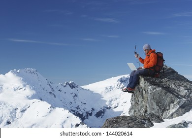 Mountain climber using laptop with walkie talkie on mountain peak against blue sky