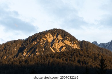 mountain covered with forest, illuminated by the warm light of the sunset. The slopes of the mountain show distinct rocky outcrops, and dense vegetation can be seen at the top. The sky is covered with - Powered by Shutterstock