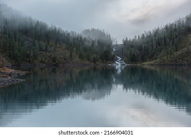 Mountain creek flows from forest hills into glacial lake in mysterious fog. Small river and coniferous trees reflected in calm alpine lake in early morning. Tranquil misty scenery with mountain lake. - Powered by Shutterstock