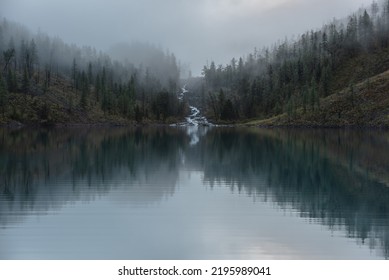 Mountain Creek Flows From Forest Hills Into Glacial Lake In Mysterious Fog. Small River And Coniferous Trees Reflected In Calm Alpine Lake In Early Morning. Tranquil Misty Scenery With Mountain Lake.