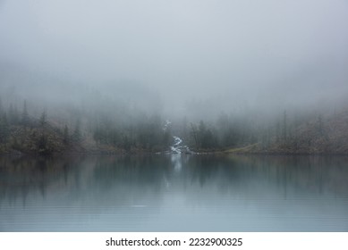 Mountain creek flows from forest hills into glacial lake in mysterious fog. Small river and coniferous trees reflected in calm alpine lake in misty morning. Tranquil landscape in fading autumn colors. - Powered by Shutterstock