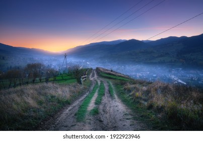 Mountain dirt road at beautiful sunset in summer. Colorful landscape with road, village in fog, blue sky with sunlight, mountains, trees at night. Trail on the hill. Travel and nature. Scenery
