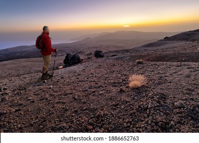 A mountain hiker with sticks stands on the highest mountain in Spain on the Atlantic island of Tenerife. He looks at the sunrise on the horizon. The clouds can be seen from above on the left. - Powered by Shutterstock