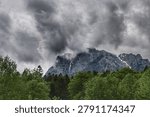 Mountain landscape with dramatic storm clouds above the Bucegi Mountains, photographed on the road to Gura Diham. Rugged rocky peaks, patches of snow, swirling mist, and a dense green forest in the foreground create a moody, atmospheric scene perfect for themes of wilderness, adventure, and dramatic weather.