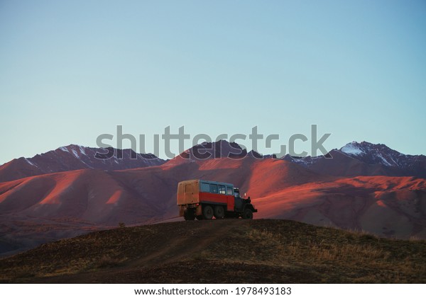 Mountain landscape with orange truck with view to\
pink magenta purple mountains with snow and sharp rocks on top\
under blue clear sky in sunset sunlight. Russia, Altai Republic, 20\
September, 2020.
