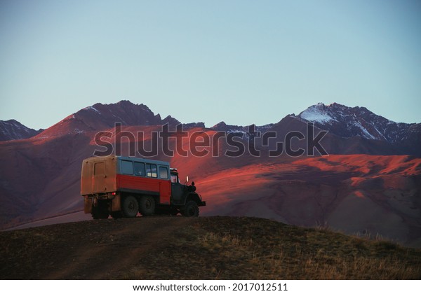 Mountain landscape with orange truck with view to\
pink magenta purple mountains with snow and sharp rocks on top\
under blue clear sky in sunset sunlight. Russia, Altai Republic, 20\
September, 2020.