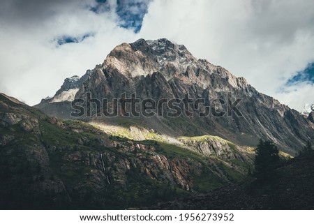 Mountain landscape with rocks with snow in sunlight and low clouds on top. Awesome rocky wall with sharp rocks in sunshine. Atmospheric mountain scenery with high rocky mountain pinnacle in clouds.