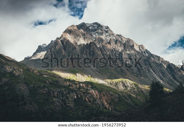 Mountain landscape with rocks with snow in\
sunlight and low clouds on top. Awesome rocky wall with sharp rocks\
in sunshine. Atmospheric mountain scenery with high rocky mountain\
pinnacle in clouds.