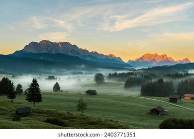 Mountain meadows with small huts in light ground fog and Zugspitz massif in the background at sunrise, Krün, Bavaria, Germany - Powered by Shutterstock