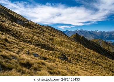 A mountain path offering scenic views of rolling hills, a tranquil lake, and distant snow-capped mountains under a partly cloudy sky. The rugged landscape and clear sky create a serene and inviting a - Powered by Shutterstock