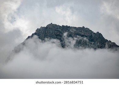 Mountain peak rises through thick cloud layers. Its rocky face shows patches of green moss. Overcast skies surround the misty slopes. The scene feels quiet and untouched