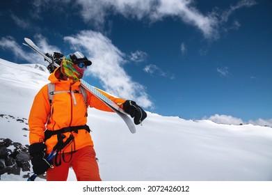 Mountain portrait of a professional freeride skier in orange clothing with ski poles and skis on his shoulders. Stands high in the mountains on a snowy slope