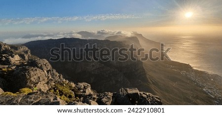 A mountain range with clouds and sun. A bright light in the sky. View of a beach and a city from a cliff. Large rock with yellow flowers.