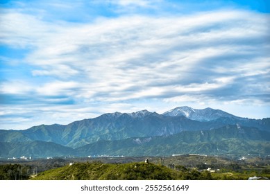 A mountain range with a cloudy sky in the background. The mountains are covered in trees. There are some houses in the distance - Powered by Shutterstock