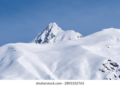 A mountain range covered with a deep layer of white snow is captured against a cloudless bright blue sky. In the center of the composition stands a pointed mountain peak. Its slopes are covered with snow with patches of exposed dark rocks that create an expressive contrast. The surface of the mountain relief is rugged with natural irregularities, depressions and ridges, which are accentuated by shadows falling on the snow.